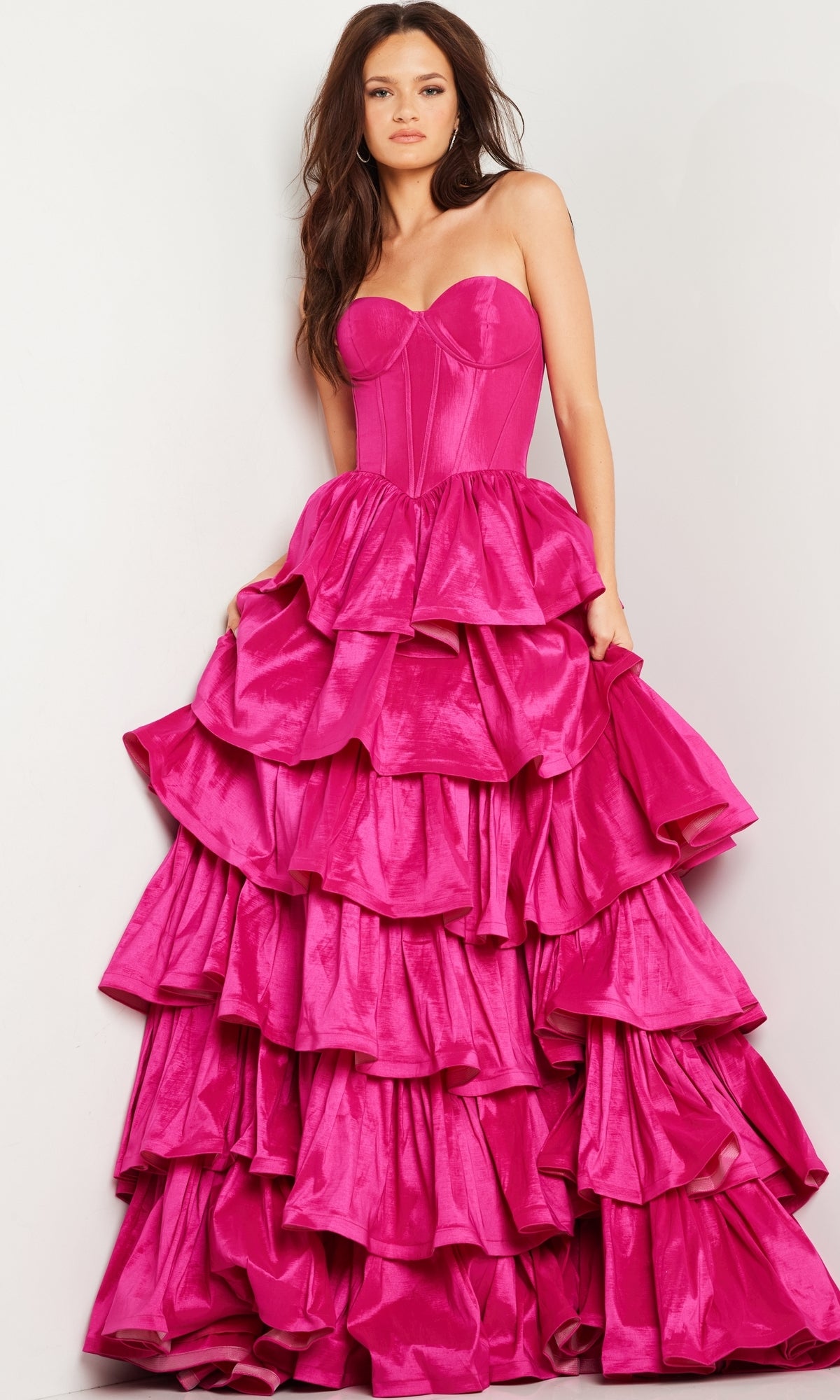 A woman wears the Strapless Ruffled Prom Ball Gown: Jovani 36619, featuring a bright pink fitted bodice and a skirt with multiple ruffled layers.